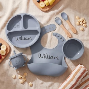 May include: A baby feeding set in a soft blue colour, including a divided plate, bowl, bib, cup, spoon, and fork. The name "William" is printed on the plate and bib. Wooden spoons are included. The set is designed for infant feeding.