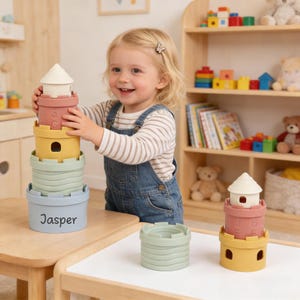 Può includere: Un bambino gioca con un set di giocattoli a castello impilabili color pastello. Il set include pezzi color pastello: azzurro, verde, giallo, rosa e bianco. La base della torre ha il nome "Jasper" stampato sopra. I giocattoli sono su un tavolo di legno.
