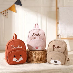 May include: Three small, personalised backpacks in orange, pink, and tan. The orange backpack has a fox face and the name "Claudia". The pink backpack has bear ears and the name "Emeru". The tan backpack has bear ears and the name "Alaric".