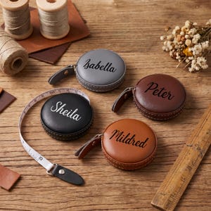 Peut inclure: Quatre mètres rubans personnalisés en similicuir gris, noir, marron et beige, chacun avec un nom différent en écriture cursive. Un mètre ruban est déployé, affichant des mesures en centimètres. Sur une surface en bois, avec des fournitures de couture.