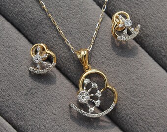 Può includere: Un set di gioielli dorati composto da collana e orecchini abbinati. La collana presenta un pendente a forma di cuore, ornato da piccole pietre scintillanti. Gli orecchini riprendono il design del pendente. Il set è presentato su una superficie grigia.