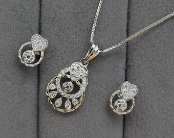 Può includere: Un set di collana e orecchini in argento. La collana presenta un ciondolo a forma di cuore e una catena. Gli orecchini hanno un design a forma di cuore. I gioielli sono su sfondo grigio.