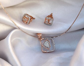 Può includere: Un set di gioielli in oro rosa con collana con pendente e orecchini abbinati. Il pendente e gli orecchini hanno una cornice quadrata con un design a forma di cuore al centro, impreziosito da piccole pietre scintillanti. Il set è appoggiato su un tessuto bianco e setoso.