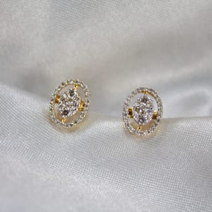 May include: A pair of gold-toned stud earrings. Each earring features a central cluster of small, clear stones surrounded by a halo of smaller, clear stones. The earrings have an oval shape and are set against a white, silky background.