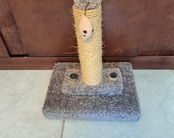 May include: A cat scratching post with a sisal-wrapped column and a plush, gray base. A small, beige mouse toy hangs from the top. The base has a square shape with a raised center section.