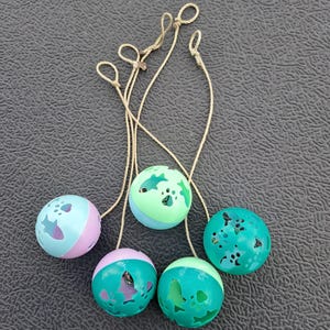 May include: A collection of five cat toys, each a hollow ball with cut-out shapes and attached string. The balls are two-toned in shades of blue, green, and pink. The cut-out shapes include stars, fish, and paw prints.