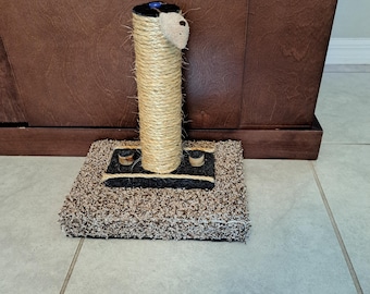 May include: A cat scratching post with a beige sisal-wrapped column and a plush mouse toy on top. The column is mounted on a rectangular base covered in brown and black carpet. The base has two wooden accents.