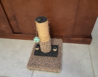 May include: A cat scratching post with a sisal rope scratching surface and a plush base. A small, green and white ball hangs from the top. The base is brown and black.