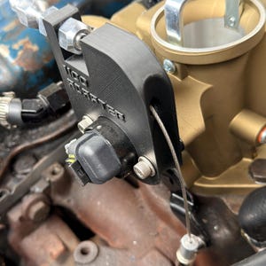 May include: A black 3D-printed throttle position sensor bracket with the text "1100 ROASTER" embossed on the side. The component is mounted to a gold-colored throttle body and features a silver metal cable attached to a small cylindrical fitting.