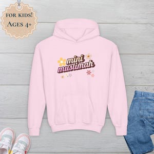 May include: A light pink hooded sweatshirt with the words "mini muslimah" in a retro font, decorated with small floral graphics. The sweatshirt has a front pocket and a drawstring hood. The tag says "For Kids! Ages 4+".