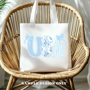 May include: White tote bag with the word "USA" in light blue. The "U" is polka-dotted, the "S" has a floral pattern, and the "A" has vertical stripes. Each letter has a light blue bow. The text "SAMPLE DESIGN ONLY" is at the bottom.
