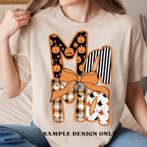 May include: Beige t-shirt with a Halloween-themed graphic. The word "Ma" is displayed in a decorative font, with the letters featuring pumpkin, spiderweb, and ghost patterns. An orange bow is centered below the letters. The text "SAMPLE DESIGN ONLY" is at the bottom.