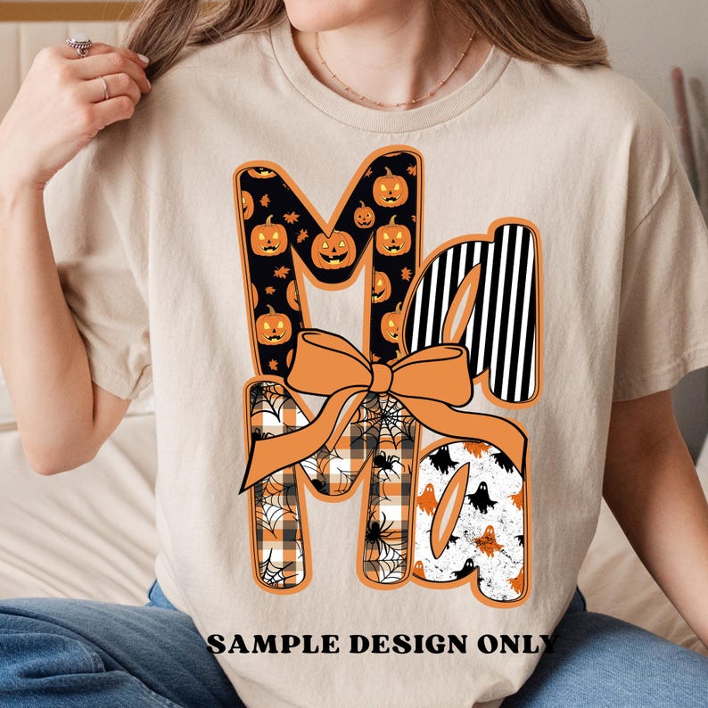 May include: Beige t-shirt with a Halloween-themed graphic. The word "Ma" is displayed in a decorative font, with the letters featuring pumpkin, spiderweb, and ghost patterns. An orange bow is centered below the letters. The text "SAMPLE DESIGN ONLY" is at the bottom.