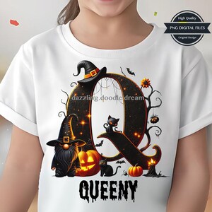 Op de afbeelding: Wit T-shirt met een Halloween-thema ontwerp. Het ontwerp bevat een grote letter "Q" met een heksenhoed, een zwarte kat, pompoenen en het woord "QUEENY" in een griezelig lettertype. Een kabouter is ook inbegrepen.
