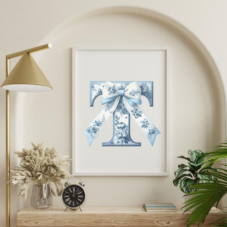 May include: A white framed print with a blue and white floral letter T with a blue bow on top. The letter is on a white background.