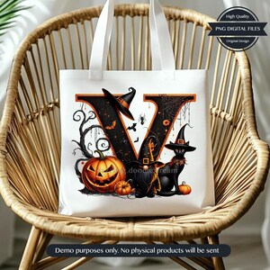 Op de afbeelding: Witte draagtas met een Halloween-ontwerp met een zwarte letter "V" versierd met een pompoen, heksenhoeden en een zwarte kat. De tas heeft ook de tekst "High Quality PNG DIGITAL FILES". De tas staat op een rieten stoel.