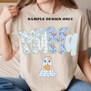 May include: Beige t-shirt with the word "Mom" in blue and white patterned letters, accented with pumpkins and a bow. A cartoon ghost holding a pumpkin is at the bottom. The text "SAMPLE DESIGN ONLY" is printed above the word "Mom".