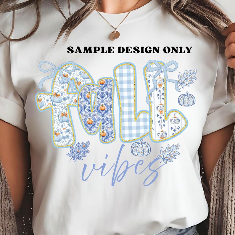 May include: White t-shirt with a "FALL VIBES" design. The word "FALL" is in a blue and white pattern with pumpkins and bows. The word "vibes" is in a light blue script. The text "SAMPLE DESIGN ONLY" is above the design.