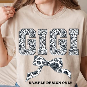 Pode incluir: Uma camiseta bege com o nome "GIGI" em letras grandes e em bloco preenchidas com um padr&atilde;o floral em preto e branco. Um gr&aacute;fico de la&ccedil;o correspondente est&aacute; abaixo do nome, com o texto "SAMPLE DESIGN ONLY" na parte inferior.