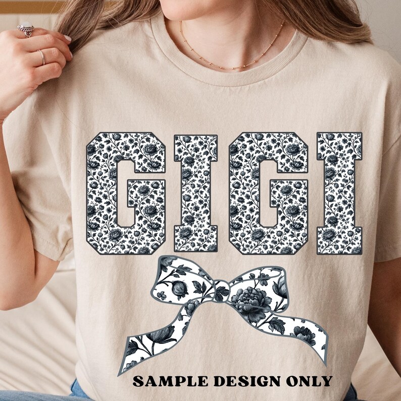 Pode incluir: Uma camiseta bege com o nome "GIGI" em letras grandes e em bloco preenchidas com um padr&atilde;o floral em preto e branco. Um gr&aacute;fico de la&ccedil;o correspondente est&aacute; abaixo do nome, com o texto "SAMPLE DESIGN ONLY" na parte inferior.
