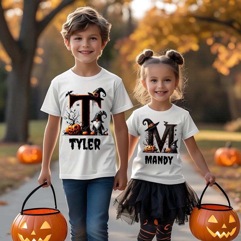 Op de afbeelding: Twee kinderen dragen witte Halloween-t-shirts met aangepaste namen en pompoenontwerpen. Het shirt van de jongen zegt "Tyler" en dat van het meisje "Mandy". Ze houden oranje pompoen-emmers vast. De achtergrond toont herfstbladeren.