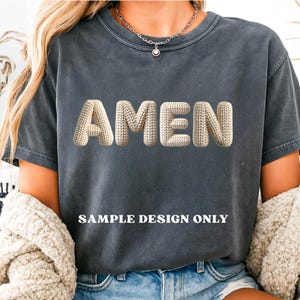 May include: Dark gray t-shirt with the word "AMEN" in a 3D knitted design. The text "SAMPLE DESIGN ONLY" is printed below the word. The shirt is a casual style.