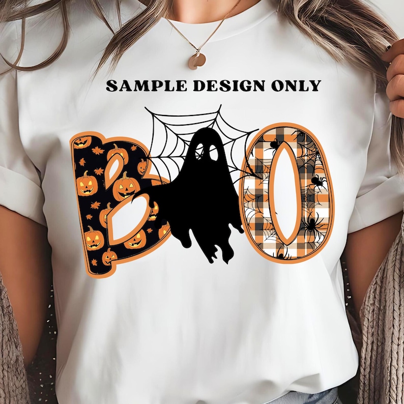 May include: White t-shirt with a Halloween-themed design. The word "BOO" is displayed with a pumpkin pattern, a black ghost, and a plaid pattern. The shirt also has a spider web and spiders.