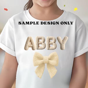May include: White t-shirt with the name "ABBY" in a beige knitted font and a beige knitted bow. The text "SAMPLE DESIGN ONLY" is printed above the name. The shirt is a simple, casual style.