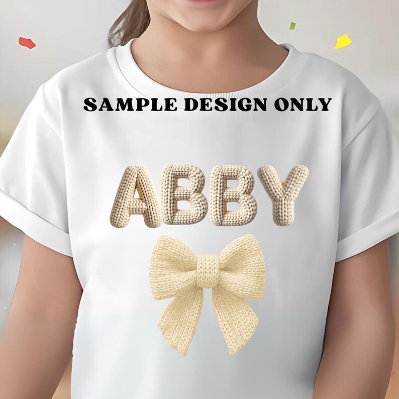 May include: White t-shirt with the name "ABBY" in a beige knitted font and a beige knitted bow. The text "SAMPLE DESIGN ONLY" is printed above the name. The shirt is a simple, casual style.