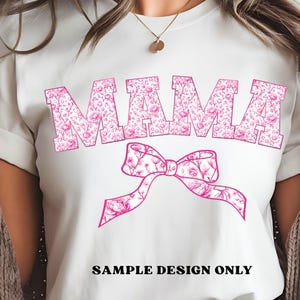 Pode incluir: Camiseta branca com a palavra "MAMA" em letras florais rosa grandes. Uma fita floral rosa est&aacute; abaixo da palavra. O texto "SAMPLE DESIGN ONLY" est&aacute; impresso em preto na parte inferior da camiseta.