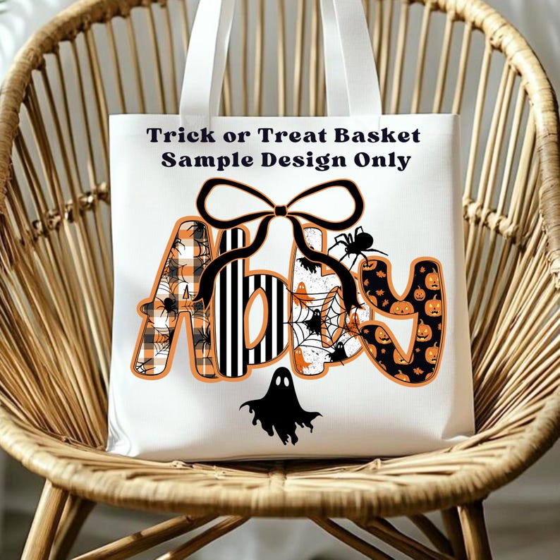 May include: White canvas tote bag with the text "Trick or Treat Basket Sample Design Only." The name "Abby" is displayed in orange and black Halloween-themed patterns, including spiders, pumpkins, and plaid. A black bow and ghost complete the design.