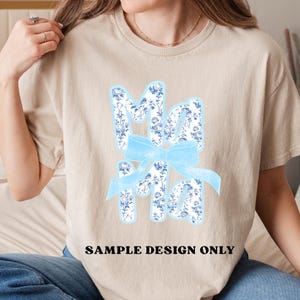 May include: Beige t-shirt with a floral "Ma Ma" design in light blue and white, accented by a light blue bow. The text "SAMPLE DESIGN ONLY" is printed in black at the bottom of the shirt.