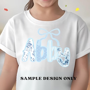May include: White t-shirt with the name "Abby" in light blue letters. The letters have floral, polka dot, and striped patterns. A light blue bow is above the name. The text "SAMPLE DESIGN ONLY" is below the name.