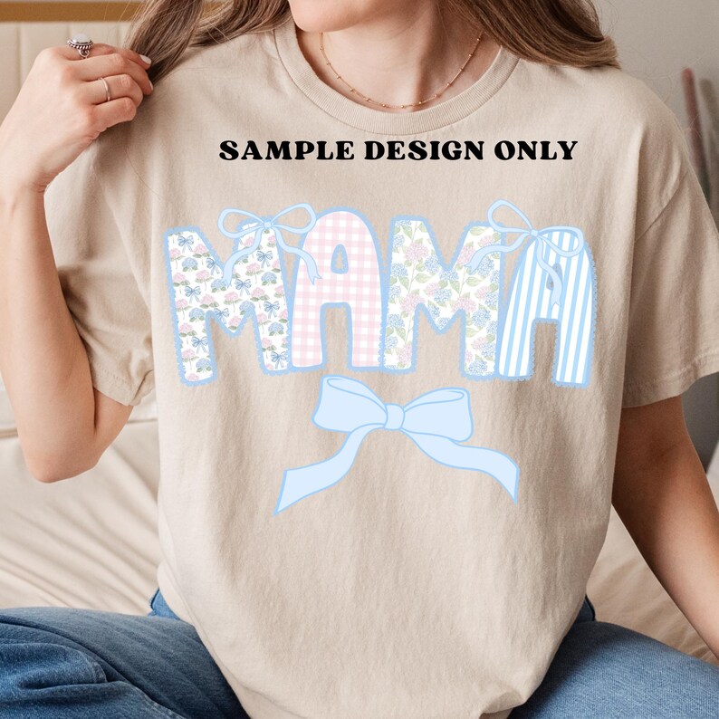 K&ouml;nnte beinhalten: Beiges T-Shirt mit dem Wort "MAMA" in dekorativer Schrift. Die Buchstaben sind mit floralen, Vichy- und gestreiften Mustern in Pastellfarben verziert, darunter eine hellblaue Schleife. Der Text "SAMPLE DESIGN ONLY" steht &uuml;ber dem Wort "MAMA".