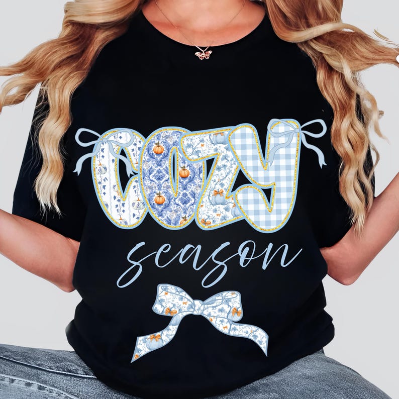 May include: Black t-shirt with the words "COZY season" in a decorative font. The word "COZY" is in a blue and white pattern with pumpkin and gingham details. A blue bow is at the bottom.
