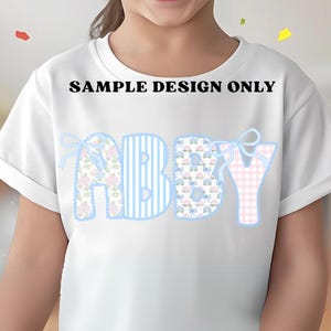 K&ouml;nnte beinhalten: Wei&szlig;es T-Shirt mit dem Namen "ABBY" in einer dekorativen Schriftart. Die Buchstaben sind mit floralen Mustern, blauen Streifen und rosa Vichy-Karo versehen. Der Text "SAMPLE DESIGN ONLY" ist &uuml;ber dem Namen aufgedruckt.