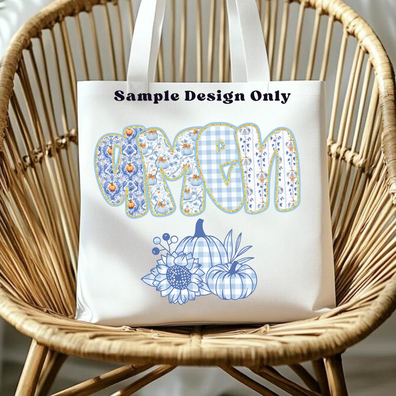 May include: White tote bag with the word "MOM" in a blue floral and gingham pattern. The bag features a blue sunflower and pumpkin design. The text "Sample Design Only" is printed at the top.