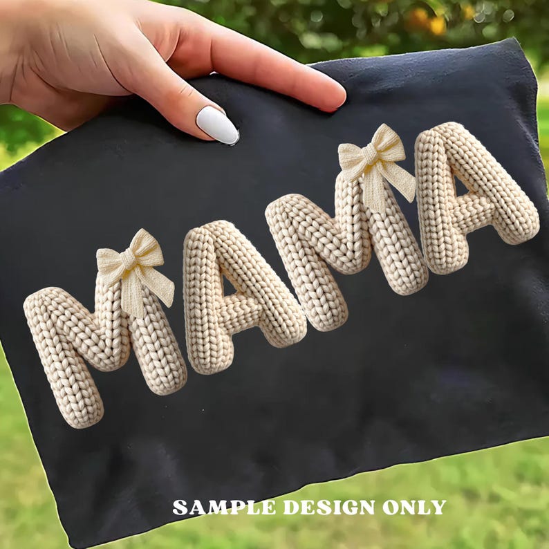 May include: Black fabric with the word "MAMA" spelled out in cream-colored, knitted letters. Each letter is adorned with a small, beige bow. The text "SAMPLE DESIGN ONLY" is printed at the bottom.