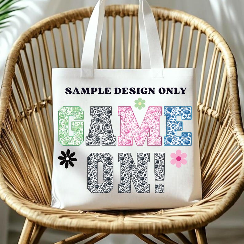 Pode incluir: Saco tote branco com o texto "SAMPLE DESIGN ONLY" acima das palavras "GAME ON!" em letras florais e estampadas coloridas. O saco tem al&ccedil;as brancas e est&aacute; numa cadeira de vime.