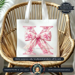 May include: A white tote bag with a pink floral letter X and a large pink bow. The letter X is surrounded by pink flowers and leaves. The tote bag is sitting on a wicker chair.