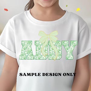 Pode incluir: Camiseta branca com o nome "ABBY" em letras verdes com padr&atilde;o floral. Um la&ccedil;o verde claro est&aacute; acima do nome. O texto "SAMPLE DESIGN ONLY" est&aacute; impresso abaixo em preto.