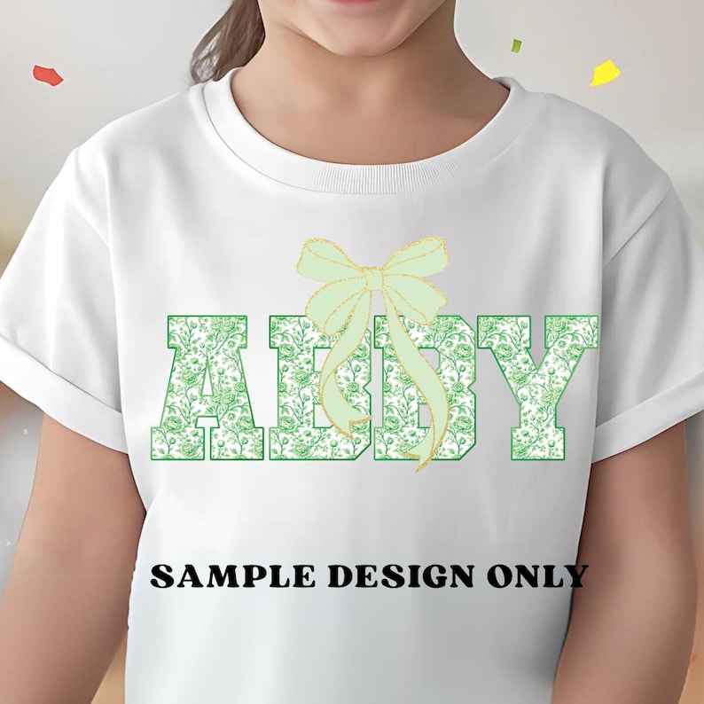 Pode incluir: Camiseta branca com o nome "ABBY" em letras verdes com padr&atilde;o floral. Um la&ccedil;o verde claro est&aacute; acima do nome. O texto "SAMPLE DESIGN ONLY" est&aacute; impresso abaixo em preto.