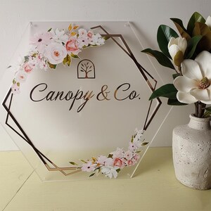 May include: Clear acrylic sign in a hexagonal shape with a gold geometric border and floral accents. The sign features the text "Canopy & Co." in a stylish font, and a tree logo. A vase with white flowers is in the background.