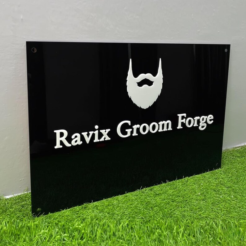 May include: A black rectangular sign with the text "Ravix Groom Forge" in white, along with a white beard graphic. The sign is mounted on a green surface, likely artificial turf. The sign has two visible mounting holes.