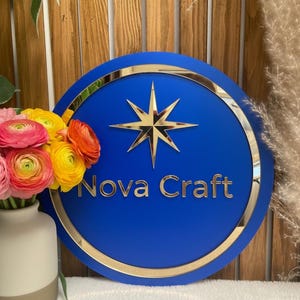 May include: A round, blue sign with gold trim and a gold starburst design. The words "Nova Craft" are in gold lettering. A vase of colorful flowers sits to the left of the sign.