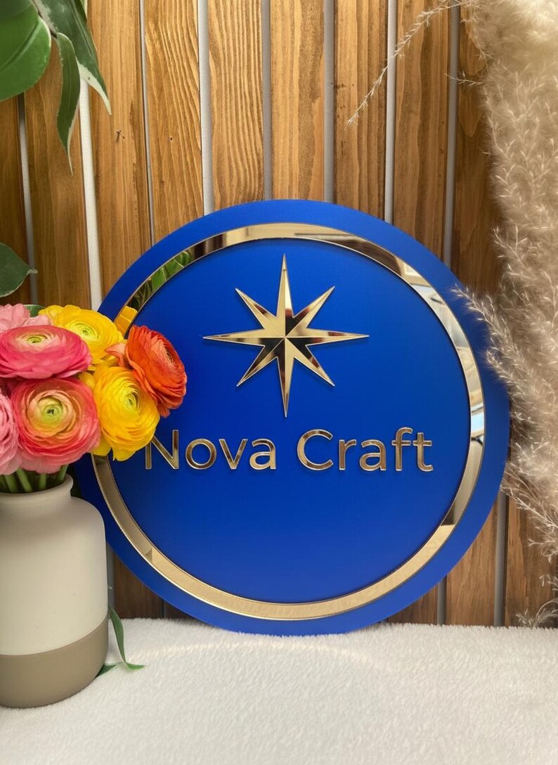 May include: A round, blue sign with gold trim and a gold starburst design. The words "Nova Craft" are in gold lettering. A vase of colorful flowers sits to the left of the sign.