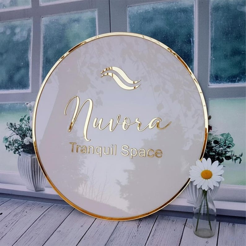 Pu&ograve; includere: Un cartello circolare con una superficie beige lucida e un bordo metallico dorato. Il cartello presenta un logo dorato con due linee curve sopra il testo Nuvora e Tranquil Space in lettere dorate. La superficie riflette la luce e il fogliame.