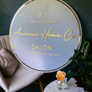 May include: A circular frosted acrylic sign with a gold metallic border. The sign features a gold leaf icon, the text "Dawnleaf symbol", "Aurinia Home Co.", "SALON", and "Bright design for everyday uplifft." in gold lettering.