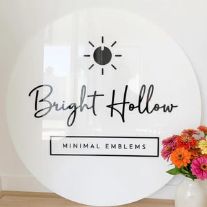 May include: A large, round white sign with the words "Bright Hollow" in black script. Above the text is a black sunburst design. Below the text is a rectangular box with the words "MINIMAL EMBLEMS". A vase of colorful flowers sits to the right.
