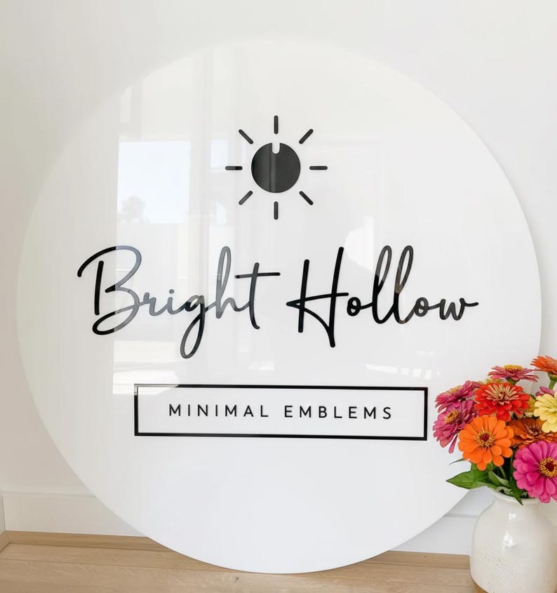 May include: A large, round white sign with the words "Bright Hollow" in black script. Above the text is a black sunburst design. Below the text is a rectangular box with the words "MINIMAL EMBLEMS". A vase of colorful flowers sits to the right.